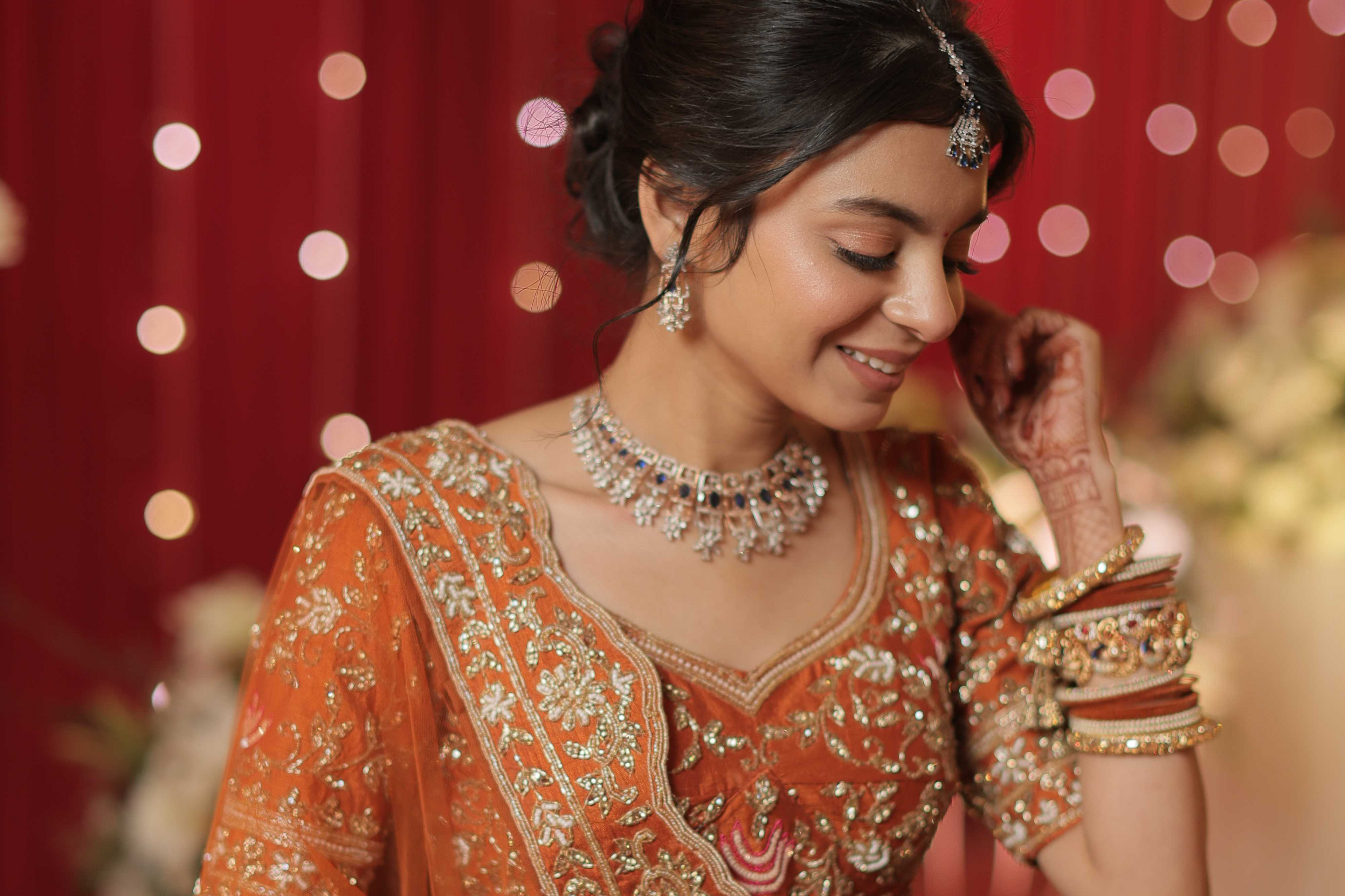 Woman in traditional orange and gold bridal outfit with jewelry against a red curtain background